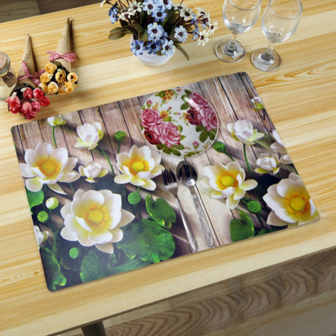 A decorative dining table mat with a white lotus flower print on a faux wood grain background, styled on a wooden table with a fork, spoon, floral saucer, and wine glasses.