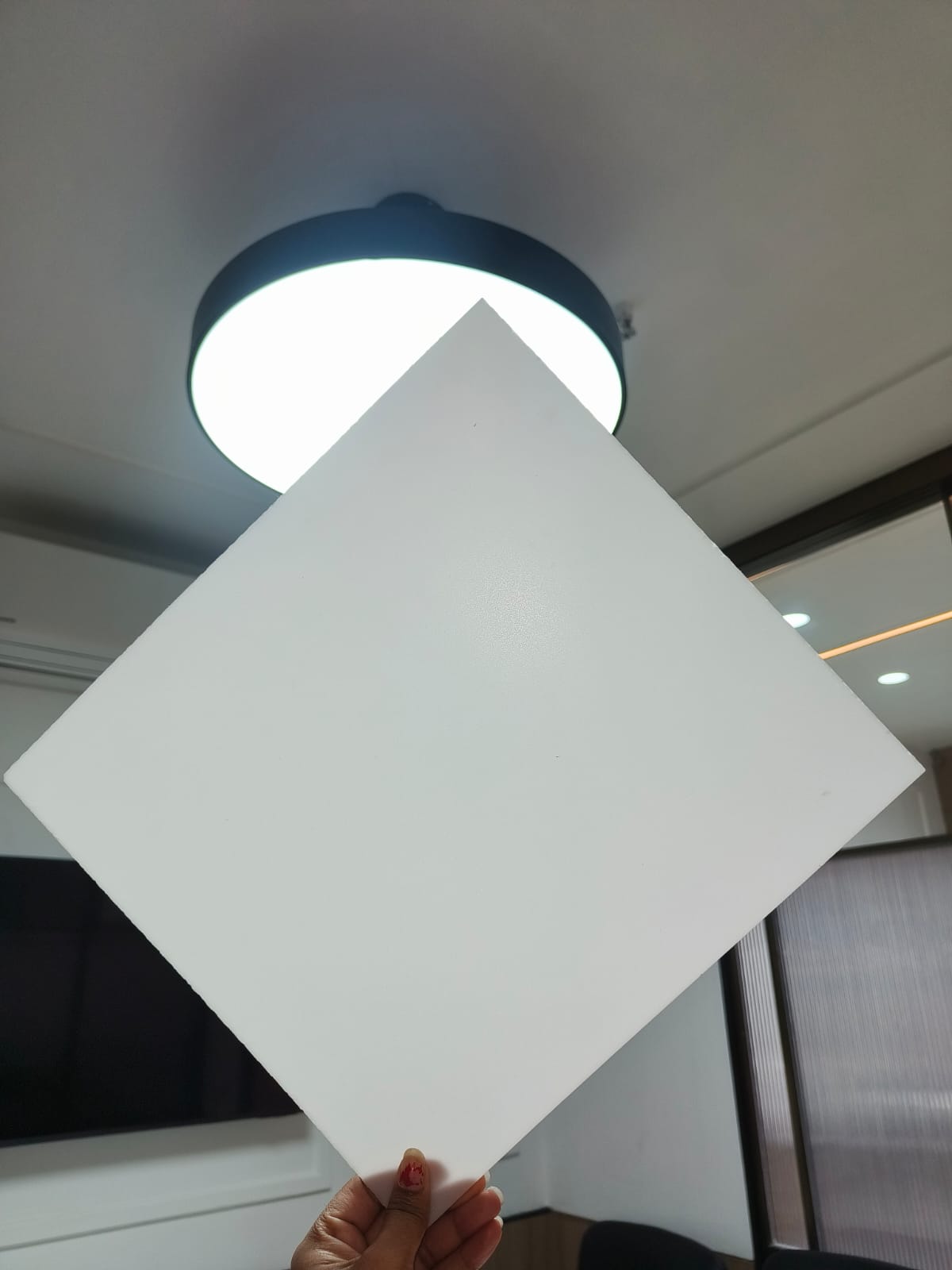 A hand holding up a square white LED diffuser sheet in front of a round ceiling light fixture.